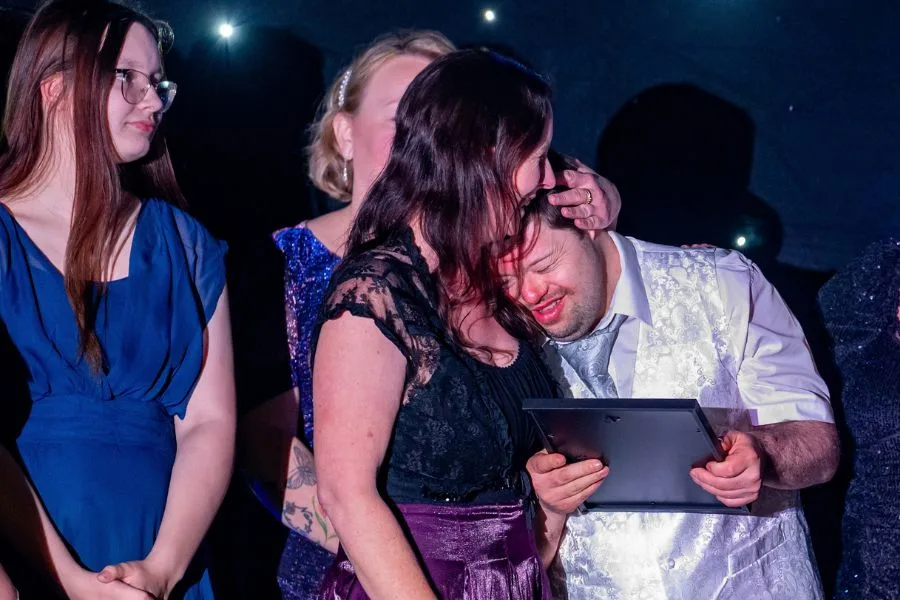 A woman embraces a man holding an award plaque on stage at the Ashford Hero Awards